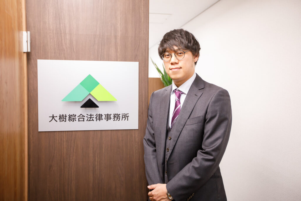 大樹綜合法律事務所の看板の前に立つ弁護士 八木翼