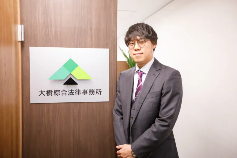 大樹綜合法律事務所の看板の前に立つ弁護士 八木翼